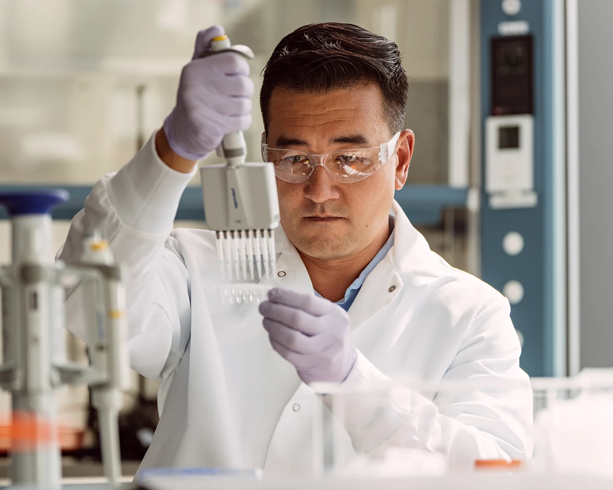 Male scientist holding an 8 lane pipette in one hand and a library tube in the other; tubes are filled with clear liquid; lab equipment in the foreground and background.